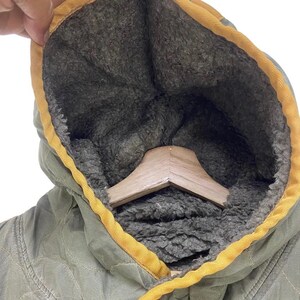 Op de afbeelding: Close-up van een olijfgroene jas met capuchon en gele rand. De binnenkant van de capuchon is gevoerd met een donkergrijs, zacht materiaal. Een houten kleerhanger is zichtbaar in de capuchon.