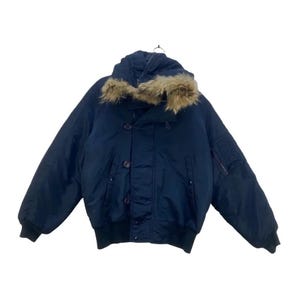 Könnte beinhalten: Marineblaue Bomberjacke mit fellbesetzter Kapuze. Die Jacke hat eine Knopfleiste, zwei Seitentaschen und eine Reißverschlusstasche am linken Ärmel. Die Bündchen und der Bund sind gerippt.