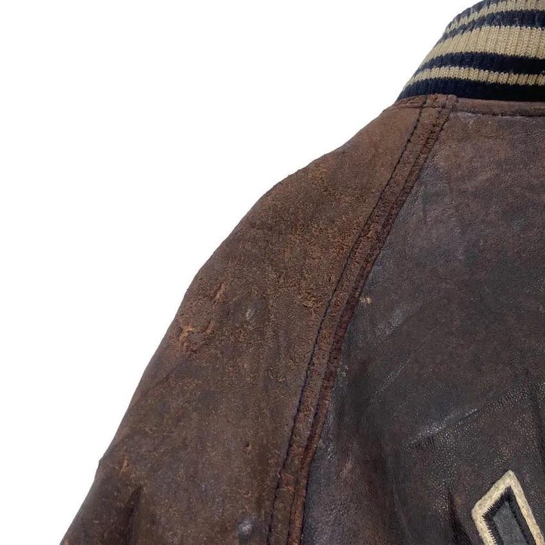 May include: Close-up of a brown leather jacket with a distressed appearance. The jacket features a ribbed collar with tan, navy, and black stripes. Visible stitching details and a partial logo add to the vintage aesthetic. The leather has a worn, textured look.