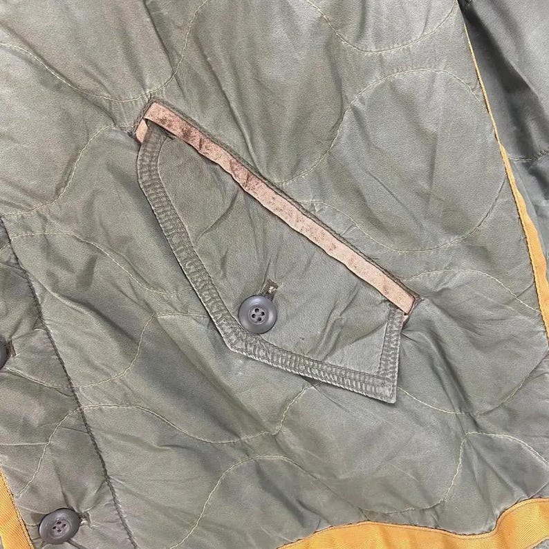 Op de afbeelding: Close-up van een gewatteerde olijfgroene jas met een zak en knoopdetails. De jas heeft een ruitpatroon en een bruine rand rond de zak. De randen van de jas zijn afgewerkt met een gele band.