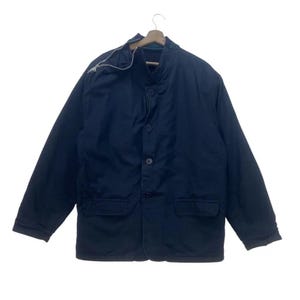 May include: A dark navy blue jacket featuring a high collar with a silver-toned zipper detail, a front button closure with four visible buttons, long sleeves, and two rectangular flap pockets positioned at the lower front of the garment.