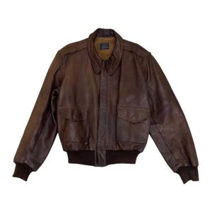 Puede incluir: Chaqueta bomber de cuero marrón con un diseño clásico. La chaqueta presenta un cuello, dos bolsillos delanteros y puños y dobladillo acanalados. El cuero tiene un aspecto desgastado, lo que sugiere un estilo vintage.