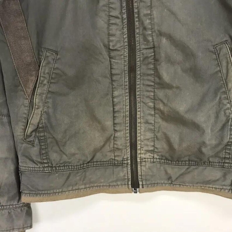 Puede incluir: Primer plano de una chaqueta verde oliva descolorida con cremallera completa. La chaqueta tiene dos bolsillos delanteros y un acento marr&oacute;n en el hombro. La tela parece desgastada, d&aacute;ndole un aspecto vintage. Prenda de vestir exterior informal.