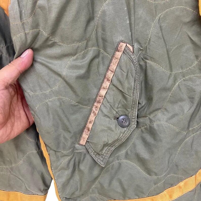 Op de afbeelding: Close-up van een gewatteerde olijfgroene jas met een bruine zak en een knoopsluiting. De jas heeft een golvend stikselpatroon en is afgewerkt met een geel-oranje rand.