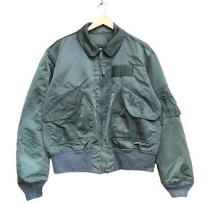 May include: A green bomber jacket with a zip-up front, two large front pockets, and a smaller pocket on the left sleeve. The jacket has a gray ribbed hem and cuffs, and a collar. The jacket is hanging on a wooden hanger.