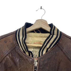 May include: A brown leather jacket with a black and beige striped collar and cuffs. The jacket features a zip closure and a label reading "American Classic". It is suspended from a wooden hanger.