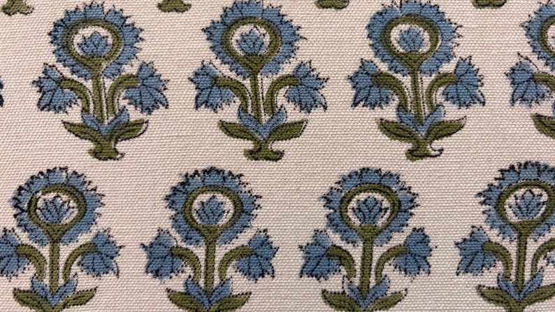 May include: Close-up of a fabric with a repeating floral pattern. The design features stylized blue flowers with green stems and leaves against a cream-colored background. The fabric has a textured appearance.