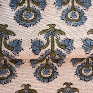 May include: Close-up of a cream-colored fabric with a repeating pattern of stylized blue and olive green floral motifs. The fabric has a textured appearance, with the floral design arranged in a symmetrical layout. The design is reminiscent of traditional block printing.