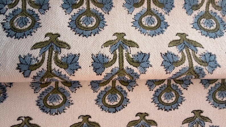 May include: Close-up of a cream-colored fabric with a repeating pattern of stylized blue and olive green floral motifs. The fabric has a textured appearance, with the floral design arranged in a symmetrical layout. The design is reminiscent of traditional block printing.