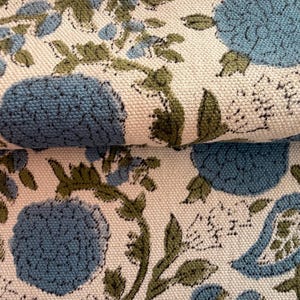 May include: Close-up of a folded fabric with a floral pattern. The design features large blue flowers, green leaves, and smaller blue floral accents on an off-white background. The fabric has a textured appearance.