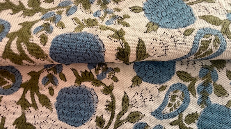 May include: Close-up of a folded fabric with a floral pattern. The design features large blue flowers, green leaves, and smaller blue floral accents on an off-white background. The fabric has a textured appearance.