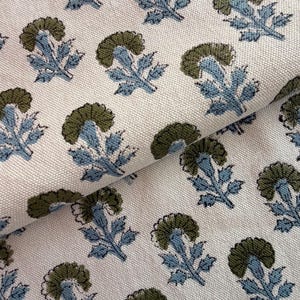 May include: Close-up of a white fabric with a repeating floral pattern. The design features stylized flowers in olive green and light blue. The fabric is folded, showing the texture and pattern.