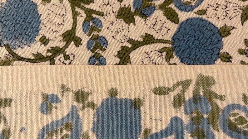 May include: Close-up of two sections of fabric. The top section features a floral pattern with blue flowers and green leaves on a cream background. The bottom section is a solid cream color with a floral pattern along the bottom edge.