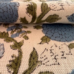 May include: Close-up of a folded fabric with a floral pattern. The design features large blue flowers and green leaves on a cream-colored background. The fabric has a textured appearance, suggesting a woven material.