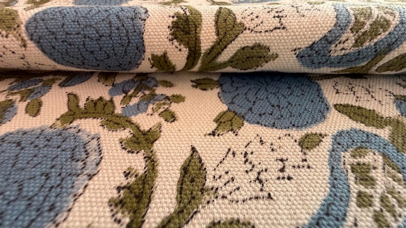 May include: Close-up of a folded fabric with a floral pattern. The design features large blue flowers and green leaves on a cream-colored background. The fabric has a textured appearance, suggesting a woven material.