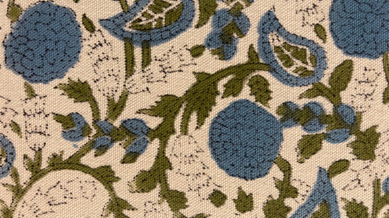 May include: Close-up of a fabric with a repeating floral pattern. The design features blue flowers and green leaves on a cream-colored background. The fabric has a textured appearance.
