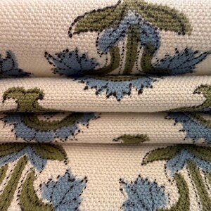May include: Close-up of folded fabric with a repeating floral pattern. The design features blue and olive green flowers on a cream-colored background. The fabric has a textured appearance, suggesting a woven material.