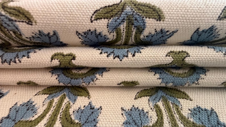 May include: Close-up of folded fabric with a repeating floral pattern. The design features blue and olive green flowers on a cream-colored background. The fabric has a textured appearance, suggesting a woven material.
