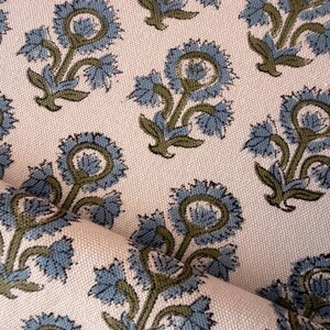 May include: Close-up of a fabric with a repeating floral pattern. The design features stylized blue flowers with green stems and leaves on a cream-colored background. The fabric is folded, showing its texture.