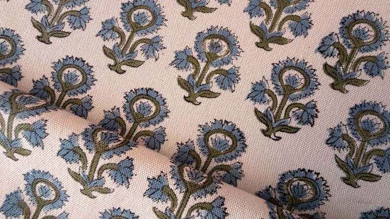 May include: Close-up of a fabric with a repeating floral pattern. The design features stylized blue flowers with green stems and leaves on a cream-colored background. The fabric is folded, showing its texture.