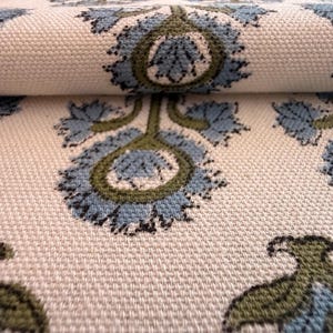 May include: Close-up of a cream-colored fabric with a repeating floral pattern. The design features blue and green stylized flowers with intricate details. The fabric is folded, revealing its texture and pattern.