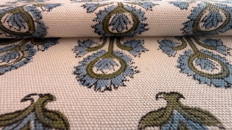 May include: Close-up of a cream-colored fabric with a repeating floral pattern. The design features blue and green stylized flowers with intricate details. The fabric is folded, revealing its texture and pattern.