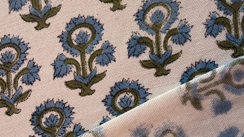 May include: Close-up of a fabric with a repeating floral pattern. The flowers are blue with green stems and leaves, set against a cream-colored background. The fabric is folded over, revealing the pattern on both sides.