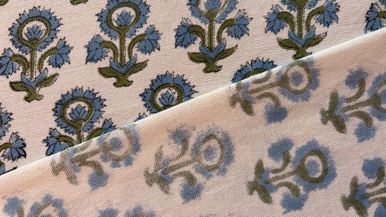 May include: Close-up of two pieces of fabric with a floral pattern. The design features blue flowers with green stems and leaves on a cream background. The fabric has a textured appearance.