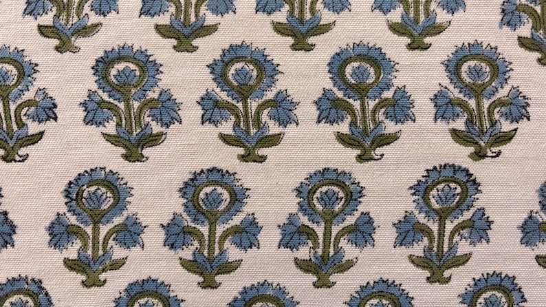 May include: Close-up of a fabric with a repeating floral pattern. The design features stylized blue flowers with green stems and leaves against a cream-colored background. The fabric has a textured appearance.