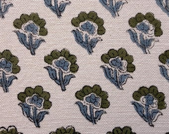 May include: Close-up of a fabric with a repeating floral pattern. The flowers have a blue center with green petal-like shapes and blue leaves. The background is a light beige color, and the fabric has a textured appearance.