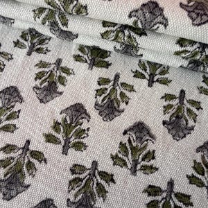 May include: Close-up of a fabric featuring a repeating pattern of stylised leaves. The leaves are in shades of grey and olive green, set against a light cream background. The fabric appears folded, showing its texture.