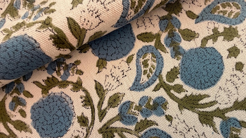 May include: Close-up of a fabric with a repeating floral pattern. The design features blue and green floral elements on a cream-colored background. The fabric is folded, showing the texture and pattern details.