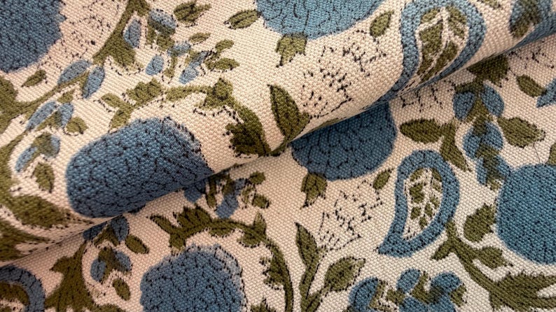 May include: Close-up of a folded fabric with a floral pattern. The design features large blue flowers and green leaves on a cream-colored background. The fabric has a textured appearance.