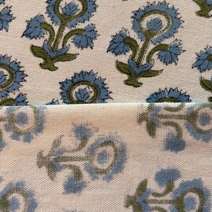 May include: Close-up of a cream-colored fabric with a repeating floral pattern. The flowers are blue with green stems and leaves. The fabric is folded over, showing the pattern on both sides.