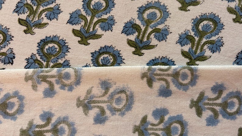 May include: Close-up of a cream-colored fabric with a repeating floral pattern. The flowers are blue with green stems and leaves. The fabric is folded over, showing the pattern on both sides.