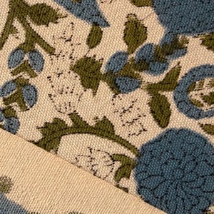May include: Close-up of a floral fabric with a cream background. The design features blue and green flowers and foliage. The fabric is folded, revealing a cream-colored edge.
