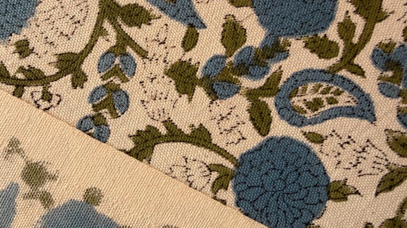 May include: Close-up of a floral fabric with a cream background. The design features blue and green flowers and foliage. The fabric is folded, revealing a cream-colored edge.
