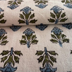 May include: Close-up of a cream-colored fabric with a repeating pattern of stylized blue and green floral motifs. The fabric is folded, revealing the texture and design. The floral pattern is evenly spaced across the material.