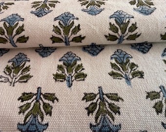 May include: Close-up of a cream-colored fabric with a repeating pattern of stylized blue and green floral motifs. The fabric is folded, revealing the texture and design. The floral pattern is evenly spaced across the material.
