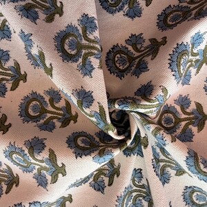 May include: Close-up of a fabric with a repeating floral pattern. The design features blue and olive green flowers on a light pink background. The fabric is folded, showing the texture and pattern.