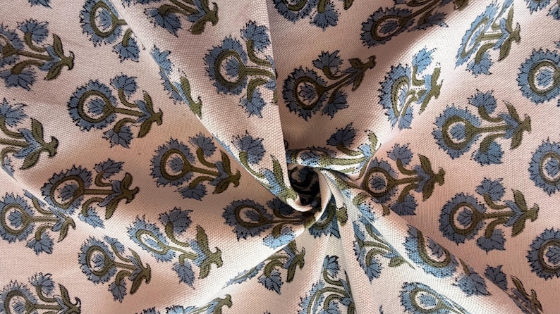 May include: Close-up of a fabric with a repeating floral pattern. The design features blue and olive green flowers on a light pink background. The fabric is folded, showing the texture and pattern.