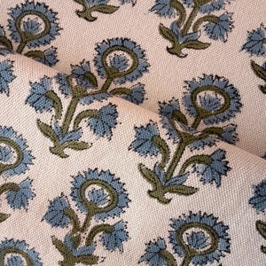 May include: Close-up of a cream-colored fabric with a repeating floral pattern. The flowers are blue with green stems and leaves. The fabric is folded, showing the texture and design.