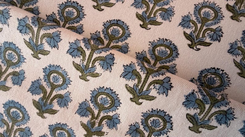 May include: Close-up of a cream-colored fabric with a repeating floral pattern. The flowers are blue with green stems and leaves. The fabric is folded, showing the texture and design.