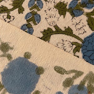 May include: Close-up of two pieces of fabric with a floral pattern. The design features blue flowers and green leaves on a cream-colored background. The fabric has a textured appearance, suggesting a natural fiber like cotton or linen.