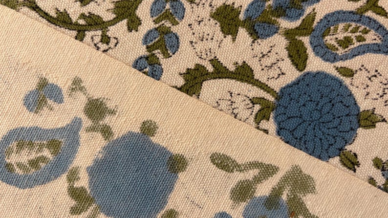 May include: Close-up of two pieces of fabric with a floral pattern. The design features blue flowers and green leaves on a cream-colored background. The fabric has a textured appearance, suggesting a natural fiber like cotton or linen.