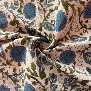 May include: Close-up of a fabric with a floral pattern. The design features large blue flowers, green leaves, and paisley shapes on a cream-colored background. The fabric is folded, showing the texture and detail of the print.