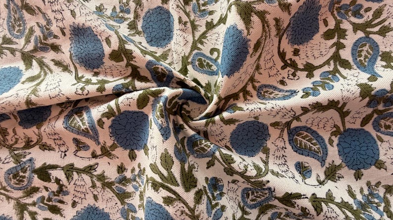 May include: Close-up of a fabric with a floral pattern. The design features large blue flowers, green leaves, and paisley shapes on a cream-colored background. The fabric is folded, showing the texture and detail of the print.