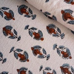May include: Close-up of a white fabric featuring a repeating floral pattern. The flowers are in rust-orange and blue-grey, with green leaves. The fabric is folded, revealing its texture and the pattern's repetition.