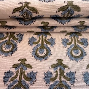 May include: Close-up of folded fabric with a repeating floral pattern. The design features blue and olive green stylized flowers on a cream-colored background. The fabric appears to be cotton or a similar woven material.
