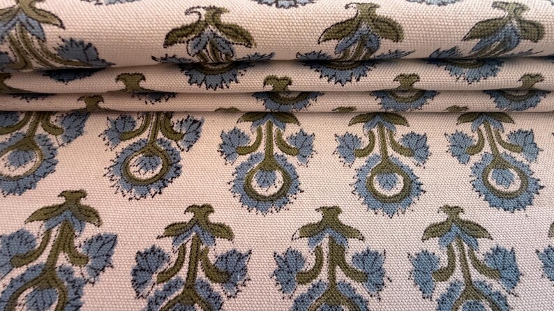 May include: Close-up of folded fabric with a repeating floral pattern. The design features blue and olive green stylized flowers on a cream-colored background. The fabric appears to be cotton or a similar woven material.